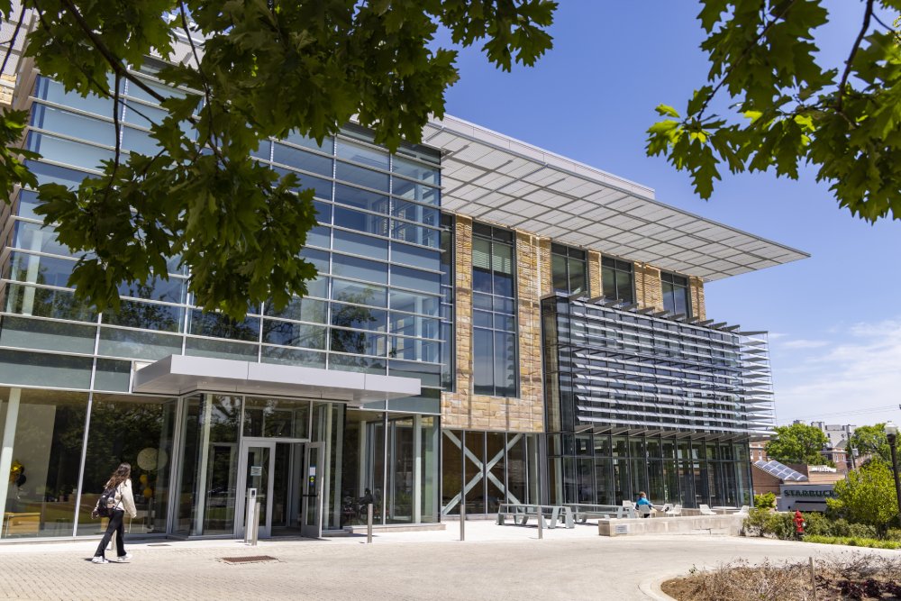 A modern glass building entrance with a metal canopy and stone accents. A person is walking toward the automatic doors, while trees and benches surround the area. In the distance, a Starbucks sign is visible.